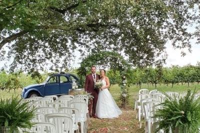 Un couple en tenue de mariage pose sous un arbre, entouré de chaises blanches et de décorations végétales en extérieur.