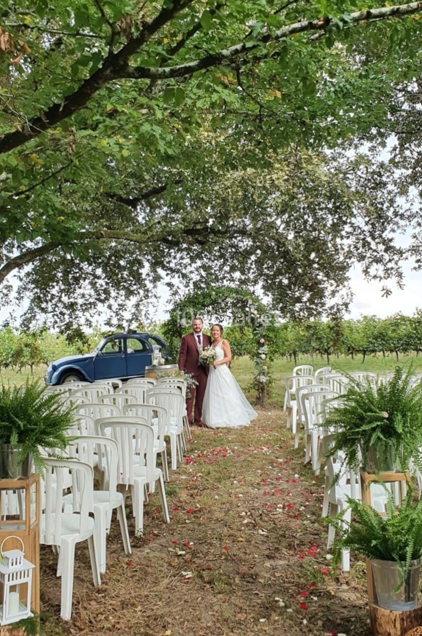 Un couple en tenue de mariage pose sous un arbre, entouré de chaises blanches et de décorations végétales en extérieur.