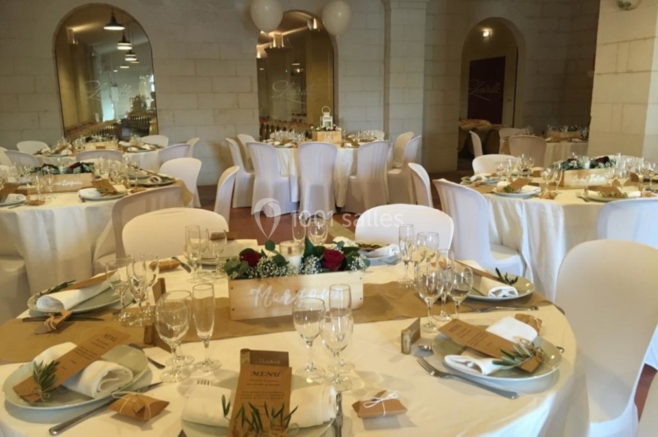 Salle de réception décorée pour un événement, avec tables rondes dressées, nappes blanches et éléments floraux.