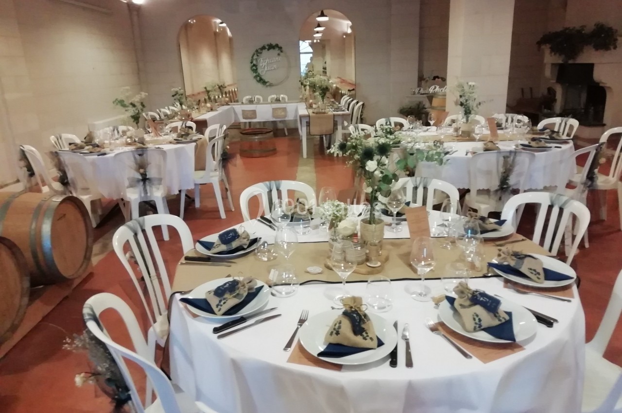 Salle de réception décorée pour un mariage, avec tables rondes dressées, nappes blanches et éléments champêtres.