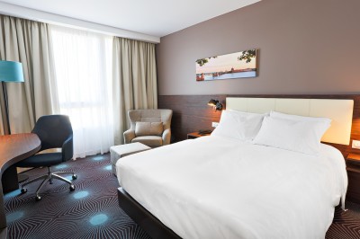 Chambre d'hôtel avec deux lits simples, un fauteuil, un bureau, une lampe et une grande fenêtre avec rideaux.