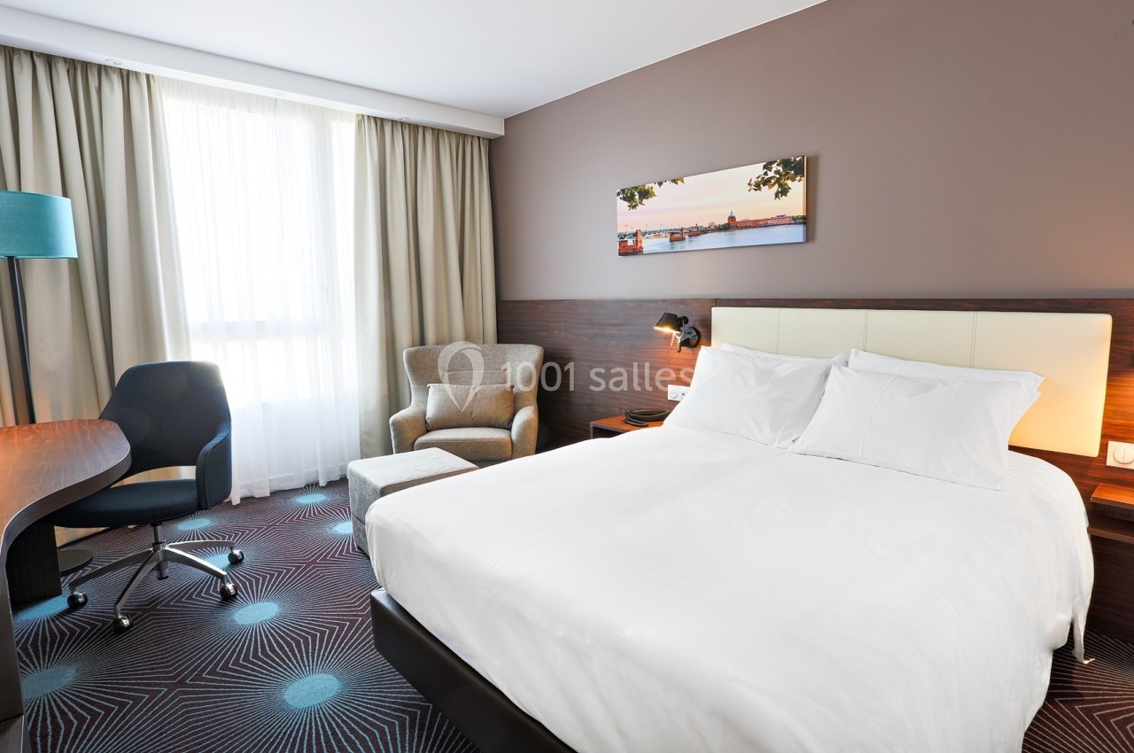 Chambre d'hôtel lumineuse avec un lit double, un fauteuil, un bureau, une chaise et une grande fenêtre avec rideaux.