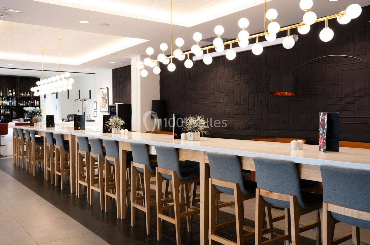 Salle moderne avec une longue table en bois, chaises rembourrées, luminaires suspendus et un mur sombre texturé.