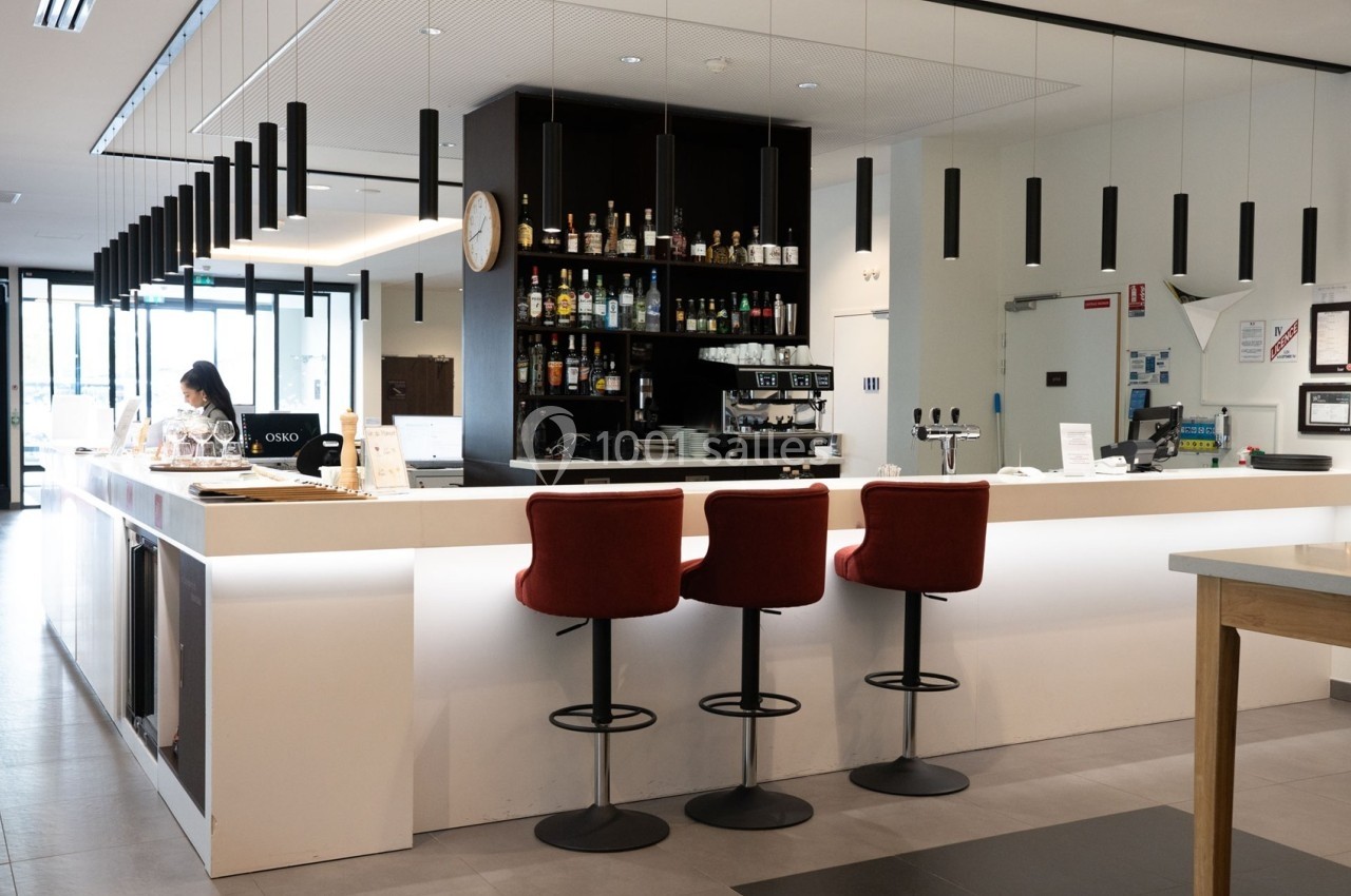 Bar moderne avec comptoir blanc, tabourets rouges, étagères de bouteilles et machine à café en arrière-plan.