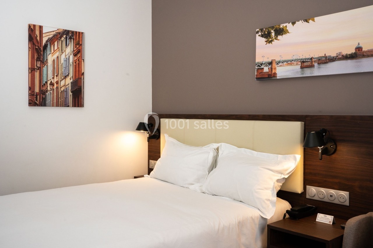 Chambre d'hôtel avec lit double, tête de lit en bois, éclairage mural et tableaux décoratifs sur les murs.