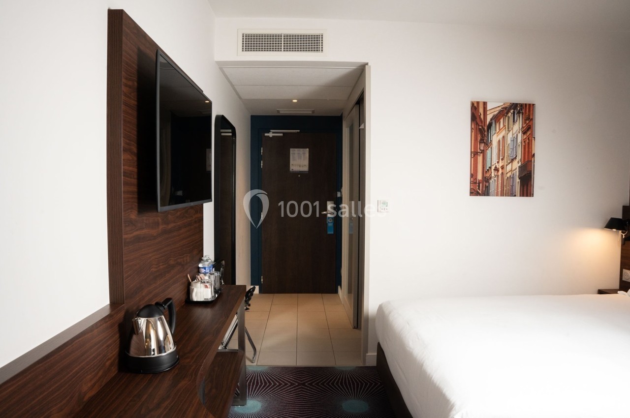 Chambre d'hôtel moderne avec lit, bureau en bois, télévision murale, bouilloire et porte d'entrée visible au fond.