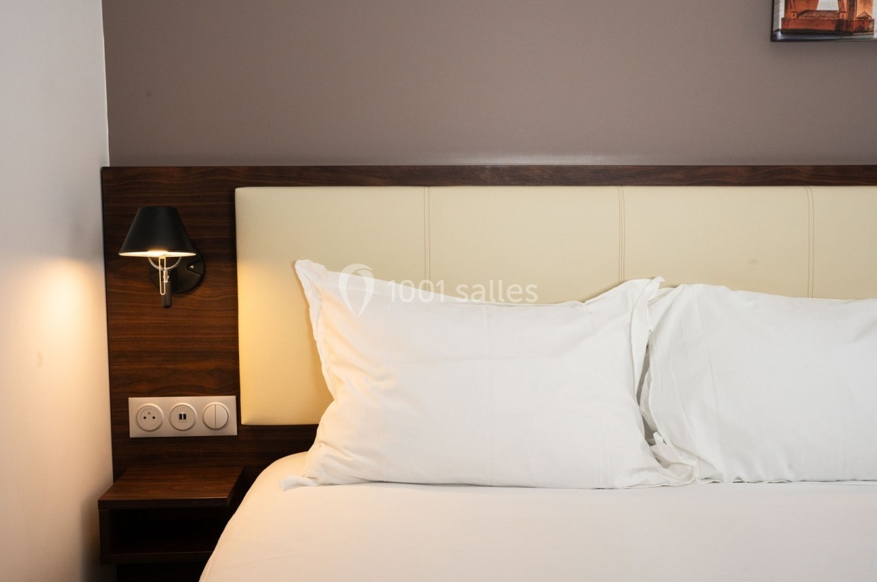 Lit avec tête de lit en bois et cuir beige, éclairé par une applique murale noire sur un mur brun.