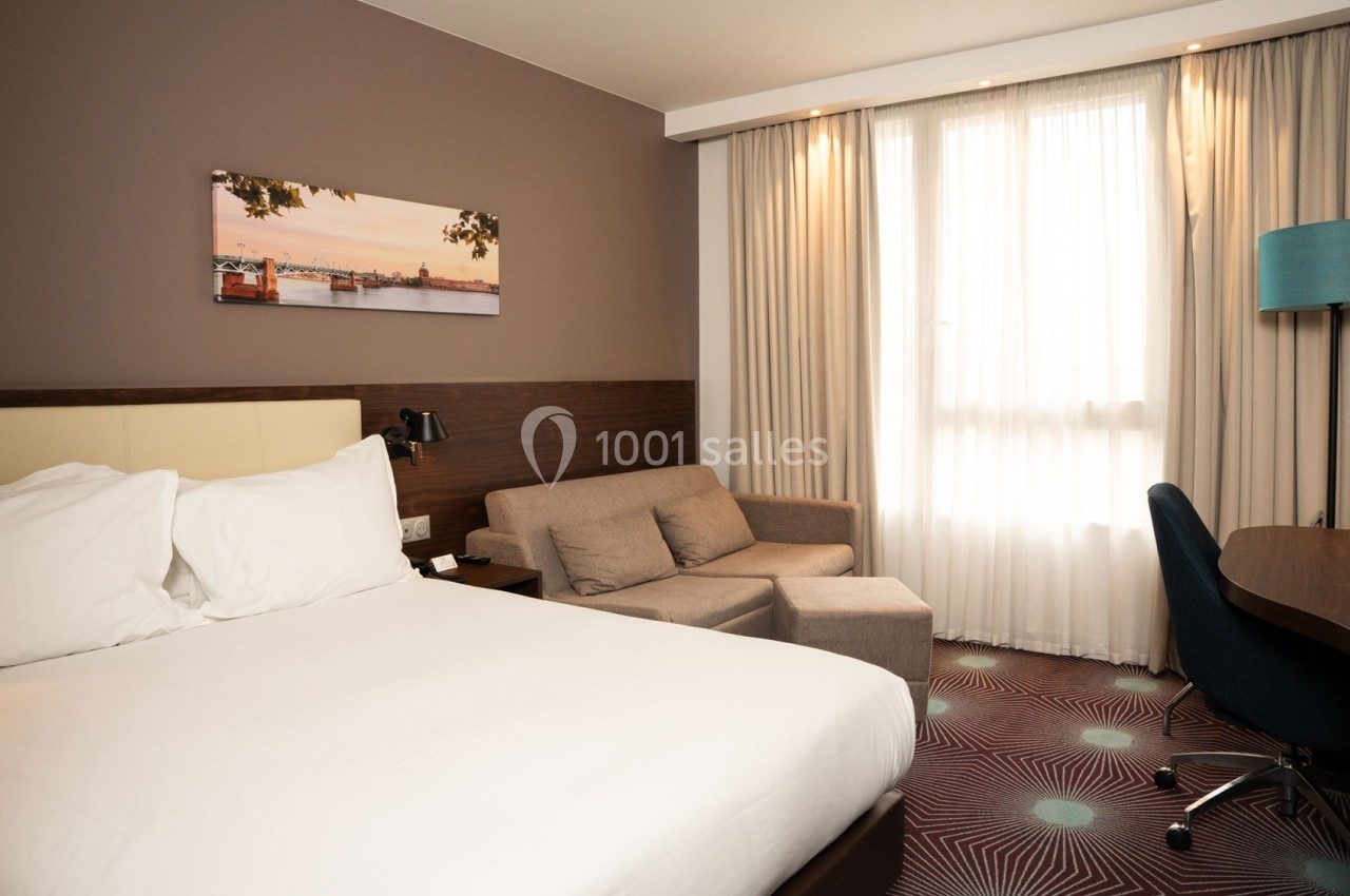 Chambre d'hôtel lumineuse avec un lit double, un canapé, un bureau et une grande fenêtre avec rideaux.