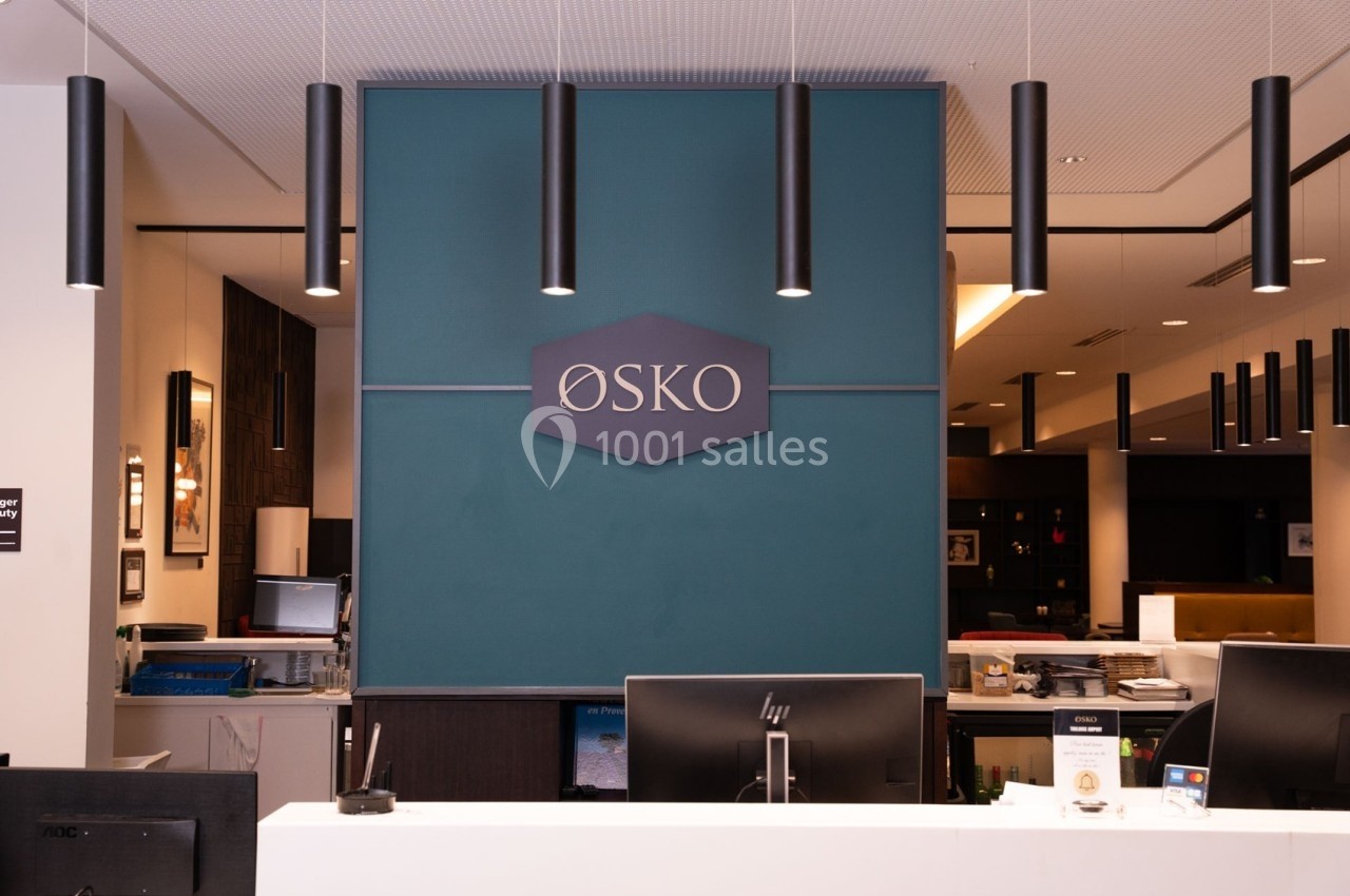 Comptoir d'accueil moderne avec un panneau ’ØSKO’ sur un mur bleu et des luminaires suspendus noirs.