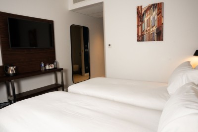 Chambre d'hôtel avec deux lits simples, un fauteuil, un bureau, une lampe et une grande fenêtre avec rideaux.