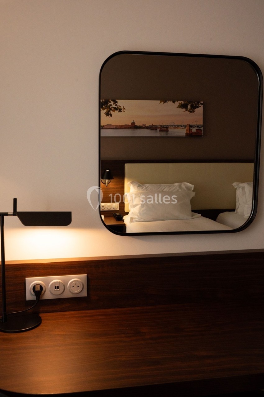 Bureau en bois avec lampe, miroir reflétant un lit et un tableau mural dans une chambre sobrement éclairée.