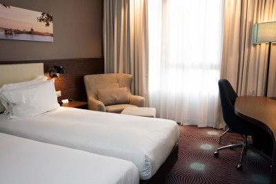 Chambre d'hôtel avec deux lits simples, un fauteuil, un bureau, une lampe et une grande fenêtre avec rideaux.