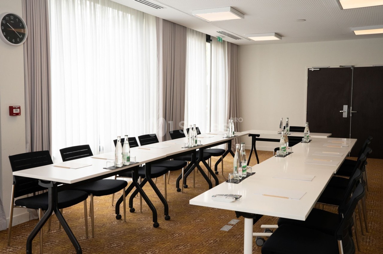 Salle de réunion lumineuse avec deux tables blanches alignées, chaises noires et bouteilles d'eau disposées.