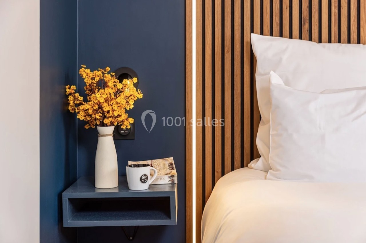 Vase avec fleurs jaunes sur une étagère bleue, à côté d'une tasse et d'un lit avec des oreillers blancs.