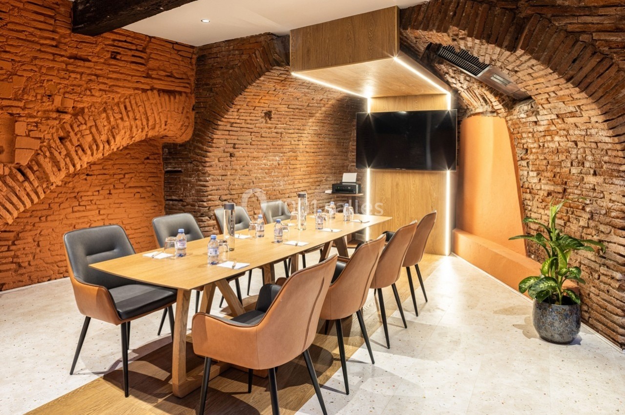 Salle de réunion avec murs en briques, table en bois, chaises modernes, écran mural et éclairage intégré.