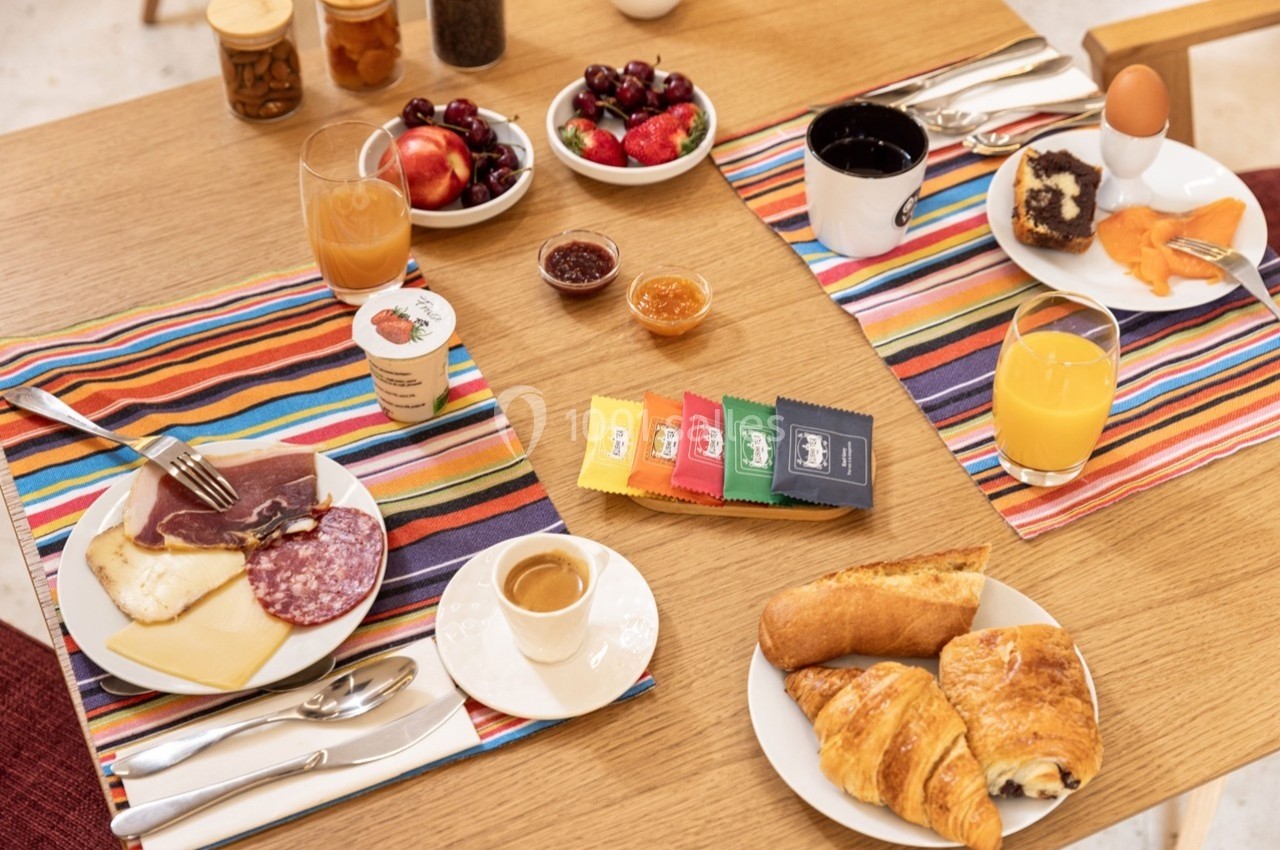 Petit-déjeuner servi sur une table avec viennoiseries, charcuterie, fromage, fruits, boissons et confitures.