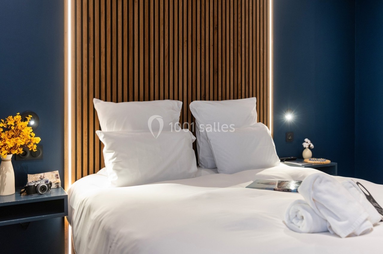 Lit double avec draps blancs, éclairage tamisé, mur en bois lamellé et tables de chevet décorées d'objets.