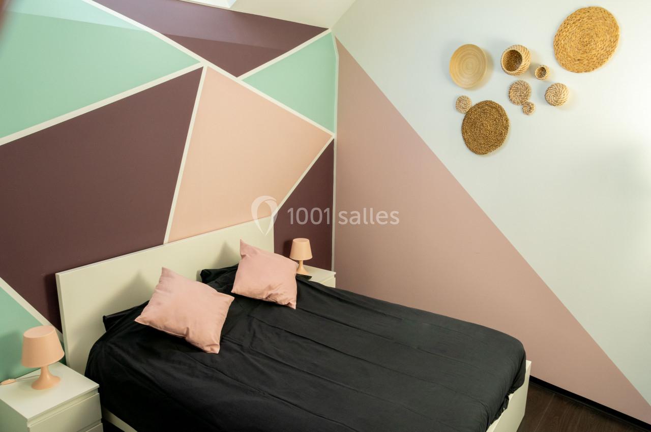 Chambre avec mur géométrique coloré, lit double noir avec coussins roses et décoration murale en osier.