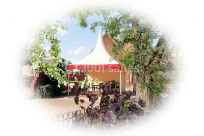 Tente blanche avec toit pointu et bordure rouge installée dans un jardin arboré, entourée de tables et chaises.