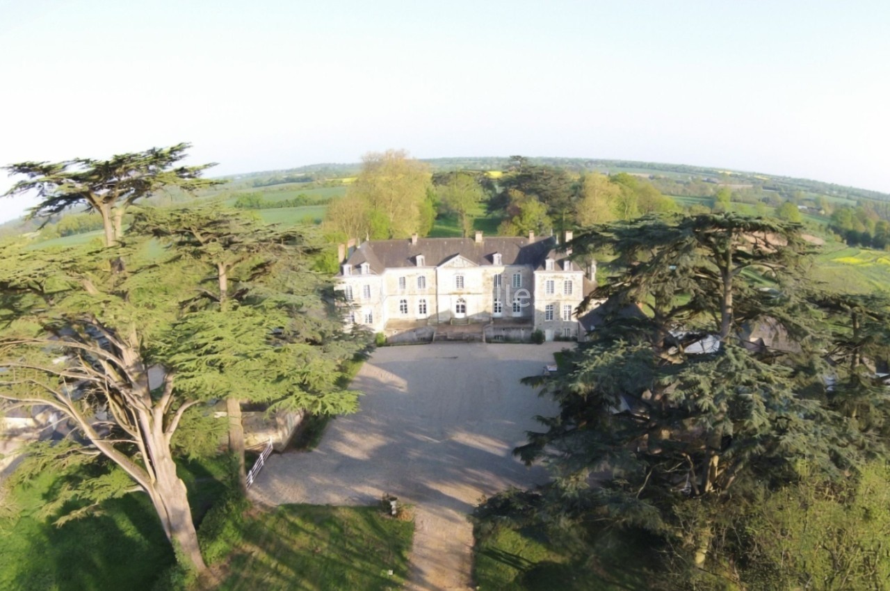 Vue aérienne d'un manoir entouré d'arbres et de champs verdoyants sous un ciel dégagé.