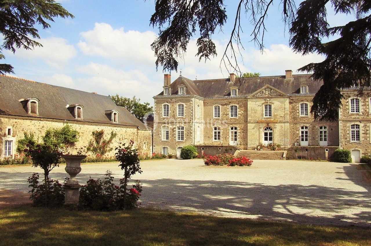 Cour d'un château en pierre avec dépendances, parterre fleuri et arbres encadrant la scène sous un ciel dégagé.