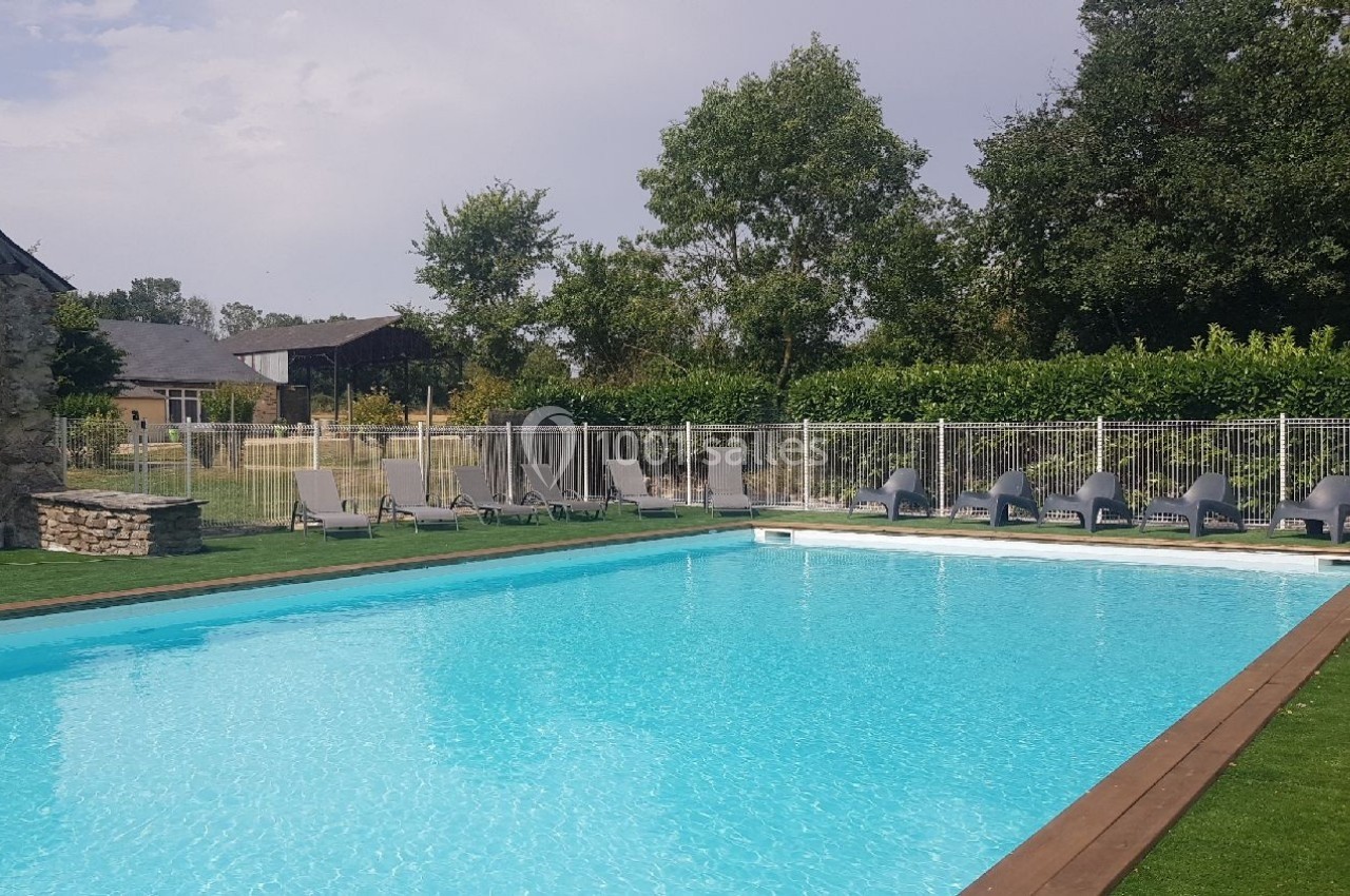 Piscine extérieure entourée de chaises longues, pelouse et arbres, avec clôture de sécurité en arrière-plan.