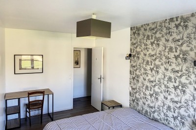 Chambre lumineuse avec lit, bureau en bois, chaise et mur décoré d'un papier peint à motifs végétaux.