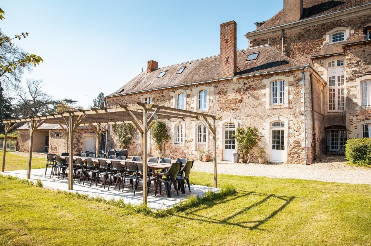 Grande table en plein air sous une pergola en bois, devant un bâtiment en pierre avec des fenêtres blanches.