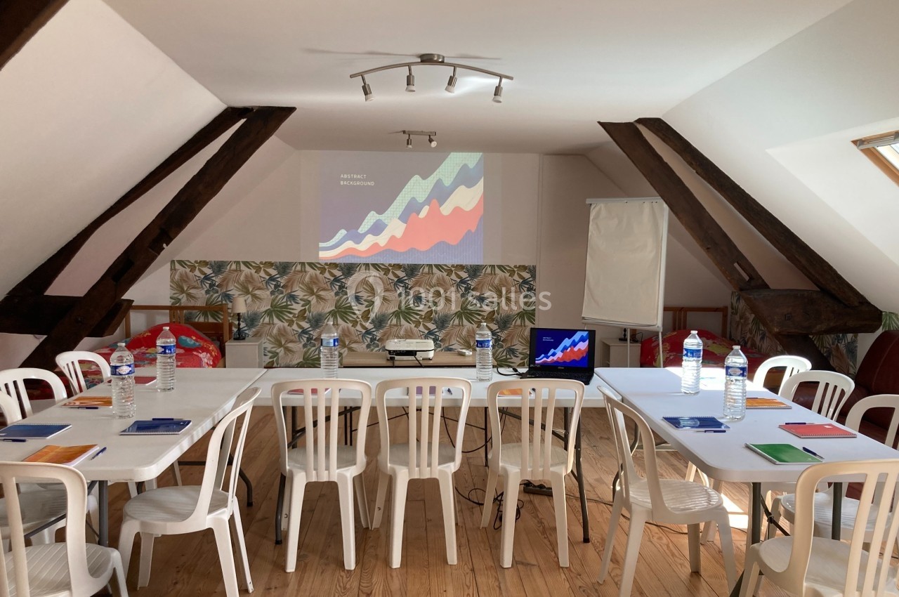 Salle de réunion lumineuse avec tables, chaises blanches, projecteur, écran, bloc-notes et bouteilles d'eau.
