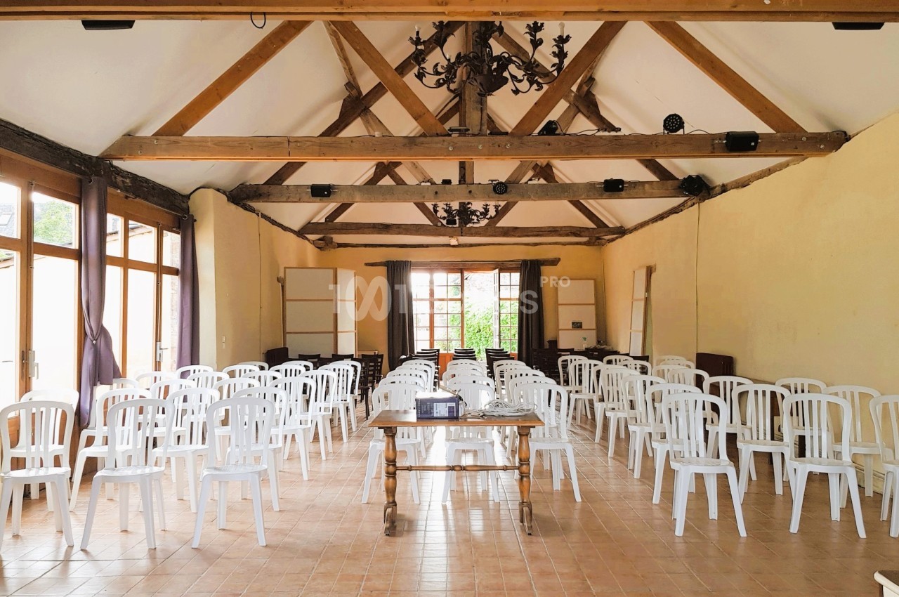 Salle lumineuse avec poutres apparentes, équipée de chaises blanches alignées et d'une table en bois au centre.