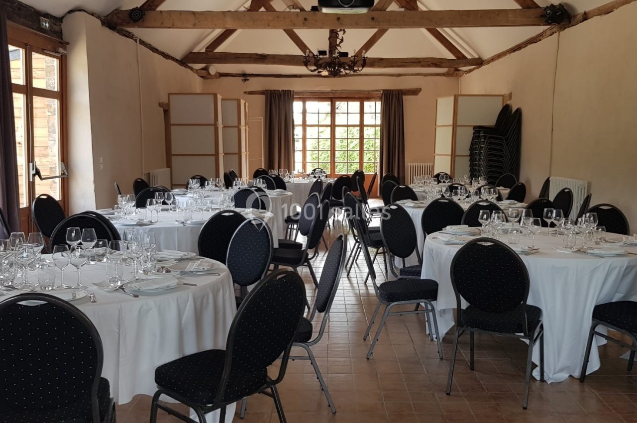 Salle de réception avec tables rondes dressées, nappes blanches et chaises noires, éclairée par des fenêtres.