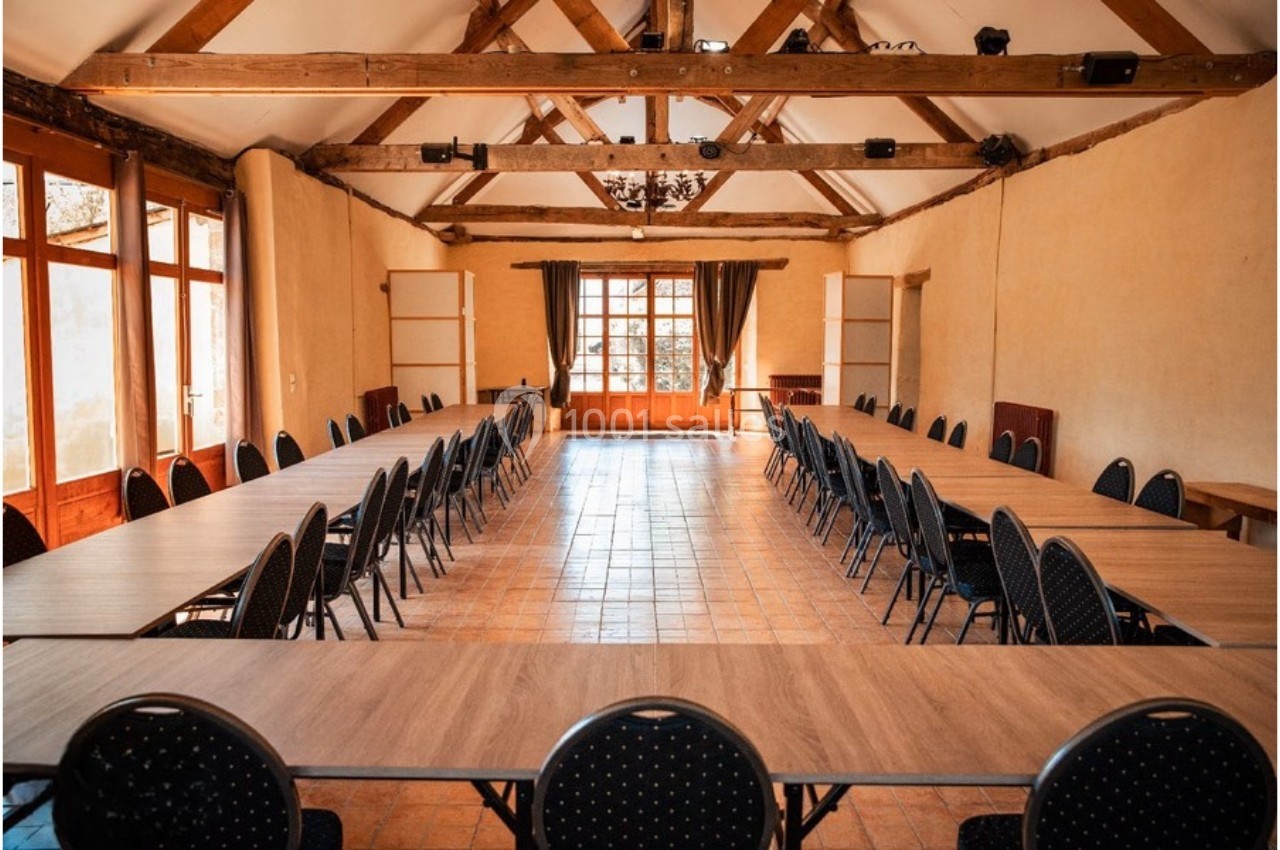Salle de réunion lumineuse avec de grandes tables en U, chaises noires et poutres apparentes au plafond.