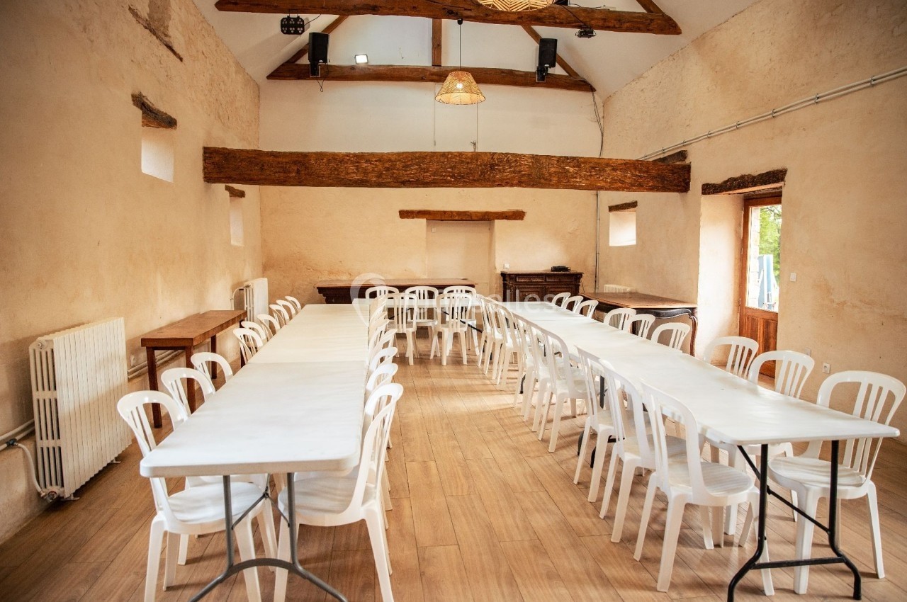 Salle lumineuse avec tables blanches alignées et chaises en plastique, poutres apparentes et sol en bois.
