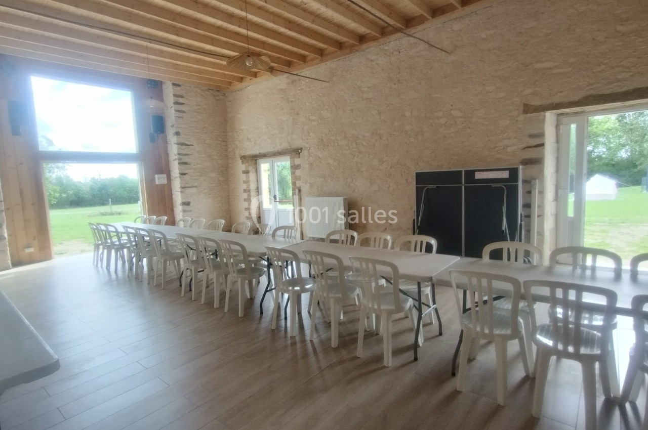 Salle lumineuse avec murs en pierre, grandes fenêtres, plafond en bois, tables et chaises alignées.