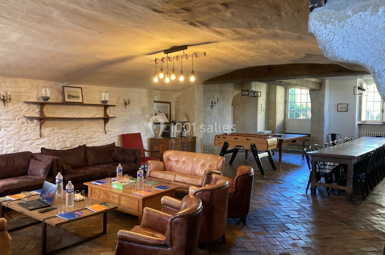 Salle voûtée avec murs en pierre, fauteuils en cuir, baby-foot, table de réunion et lumière suspendue.