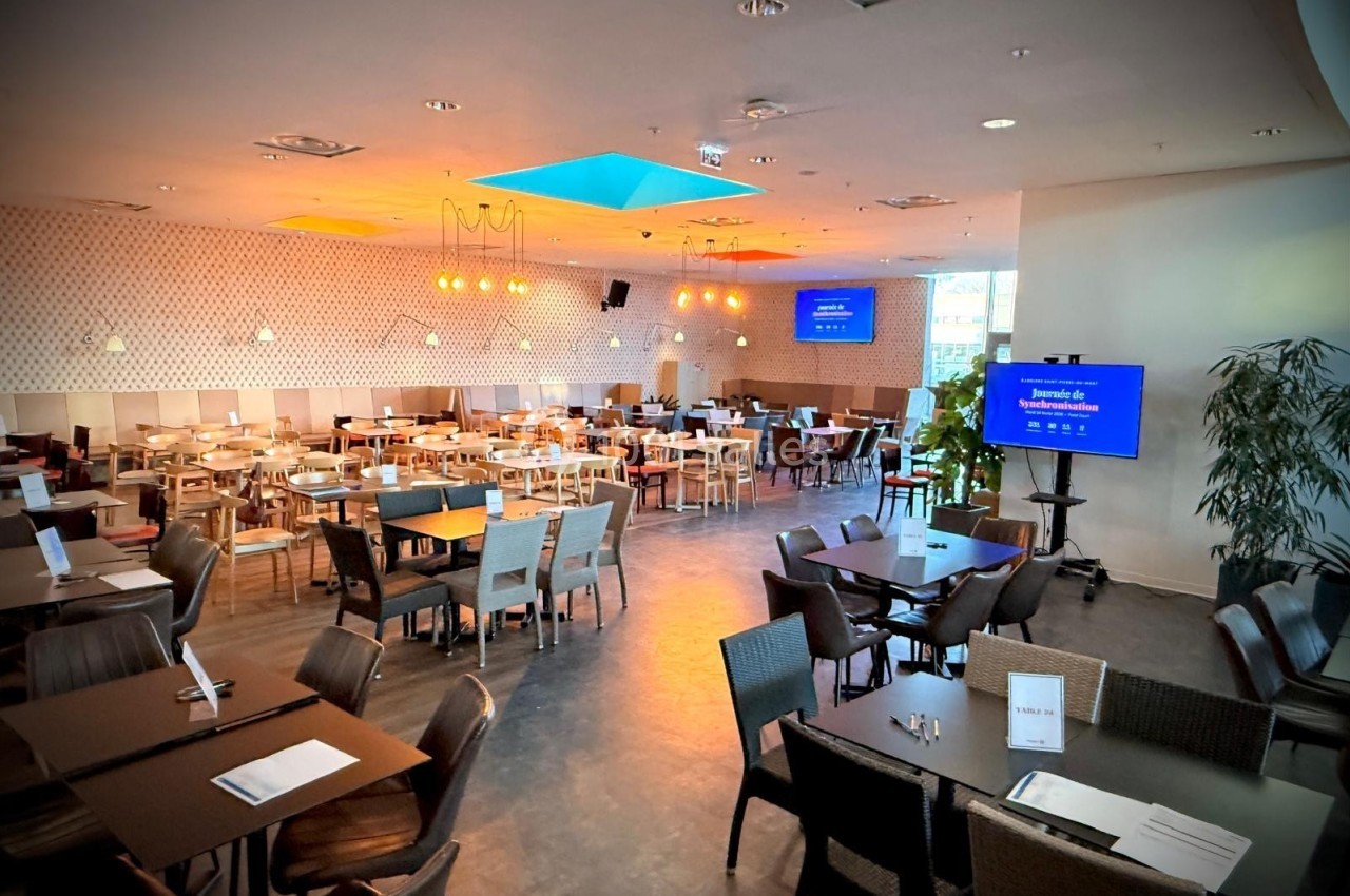 Salle de restaurant lumineuse avec tables et chaises disposées, écrans et plantes en arrière-plan.