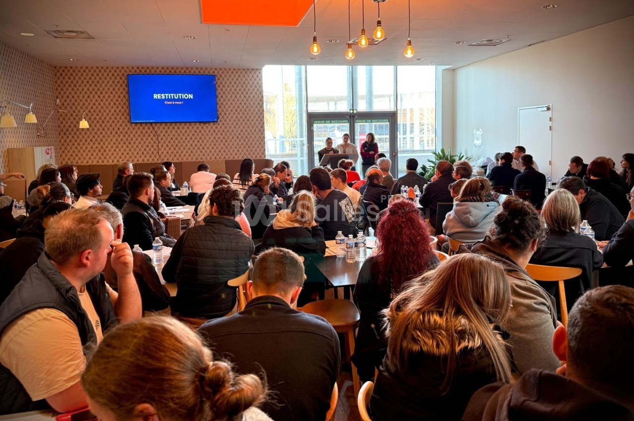 Salle comble avec des participants assis écoutant une présentation sur scène devant un écran affichant ’Restitution’.