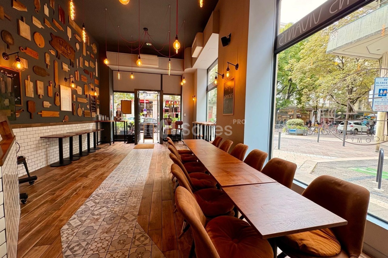 Intérieur d'un café moderne avec une grande table en bois, des chaises rembourrées et une vue sur la rue par de larges…