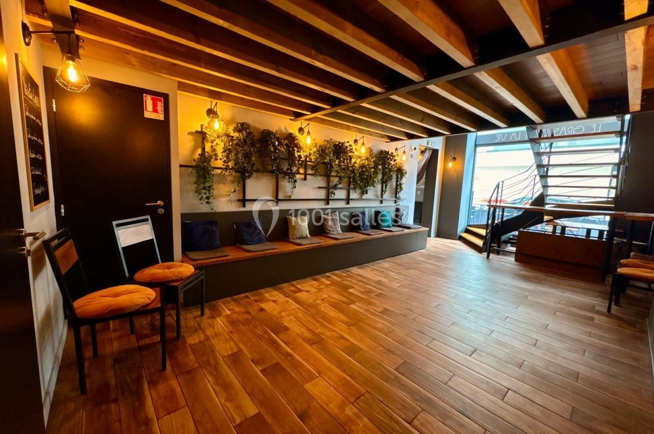 Salle lumineuse avec parquet en bois, banquette avec coussins colorés, plantes murales et plafond à poutres apparentes.