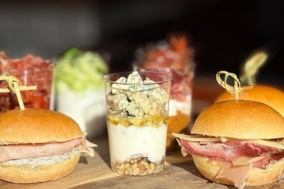Assortiment de mini-burgers au jambon et verrines garnies de fromage, noix et légumes sur une planche en bois.