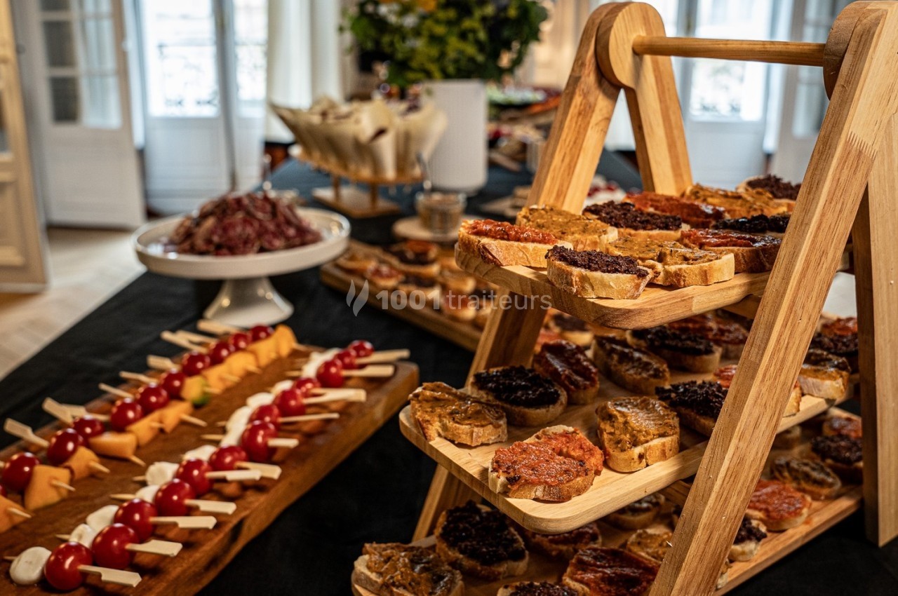 Buffet avec toasts variés sur un présentoir en bois, brochettes apéritives et plateau de charcuterie en arrière-plan.
