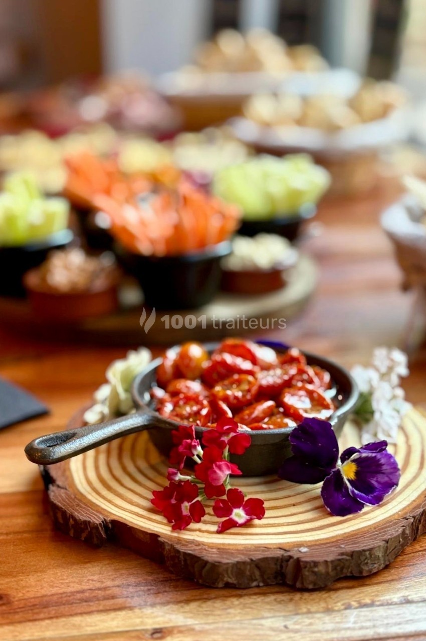 Bol de tomates cerises confites sur une planche en bois, entouré de fleurs et d'autres plats en arrière-plan flou.