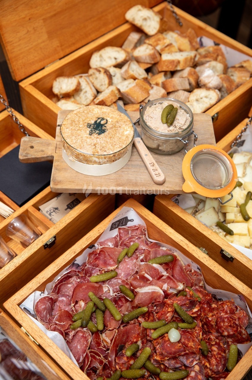 Plateau de charcuterie avec cornichons, fromage, pain tranché et rillettes présentés dans des boîtes en bois.