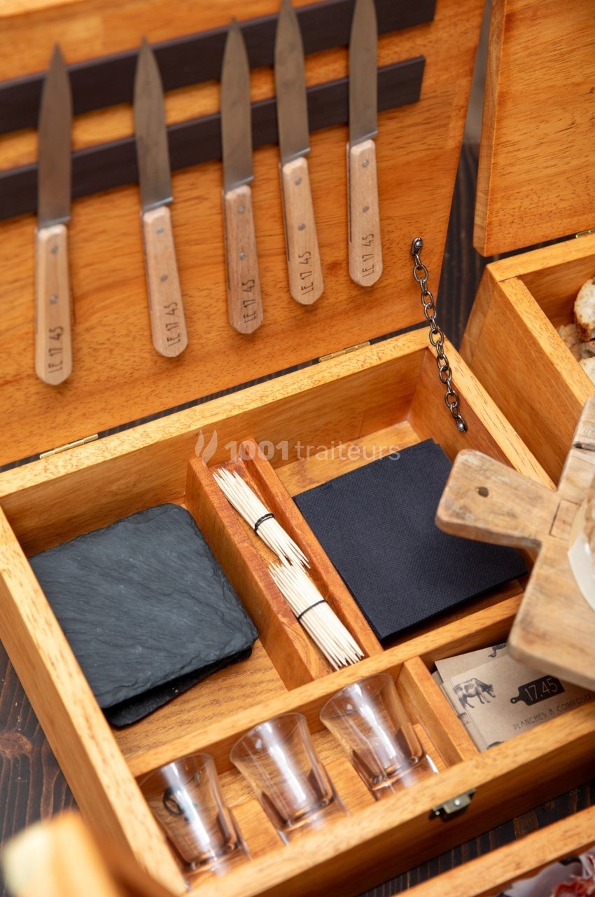 Boîte en bois ouverte contenant des couteaux, des piques, des verres, une ardoise et des serviettes noires.