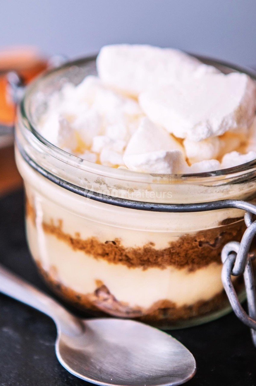 Dessert en verrine avec couches de crème, biscuit émietté et morceaux de meringue, posé sur une ardoise avec une cuillère.