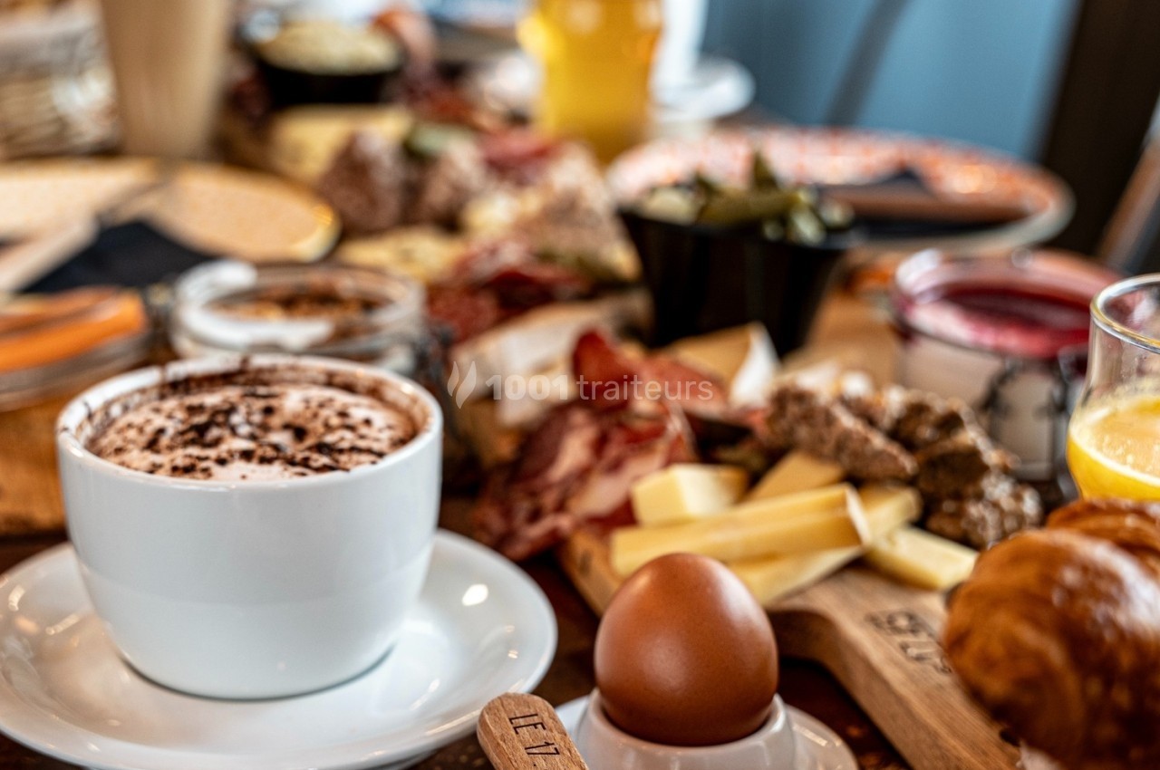 Petit-déjeuner varié avec café, œuf à la coque, viennoiseries, fromages, charcuteries et jus d'orange sur une table.