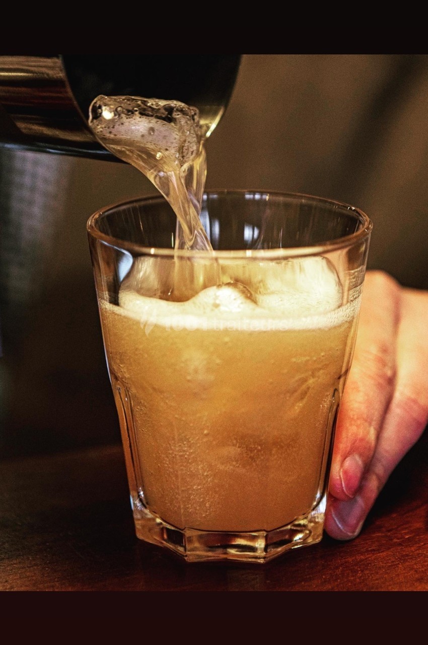 Un liquide mousseux est versé dans un verre transparent tenu par une main sur une table en bois.