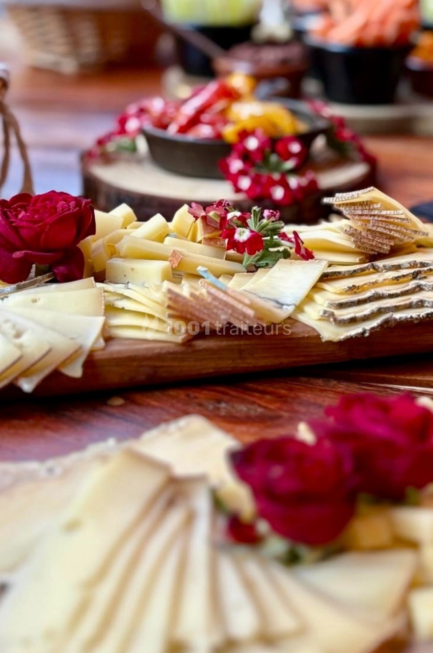 Plateau de fromages variés décoré de fleurs rouges, présenté sur une table en bois avec d'autres amuse-bouches en arrière…