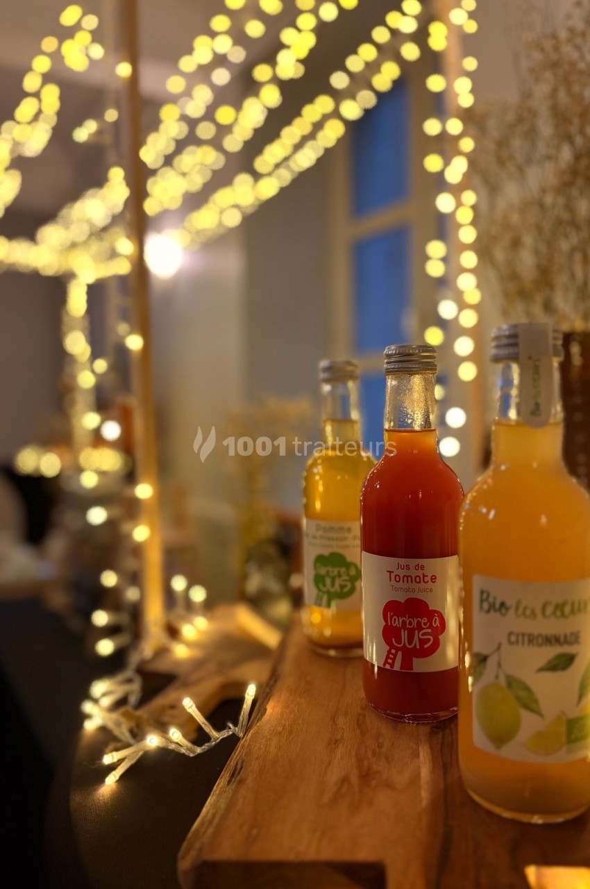 Bouteilles de jus artisanaux disposées sur un support en bois, éclairées par des guirlandes lumineuses dans un décor…