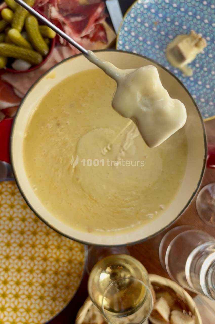Fondue au fromage dans une casserole, avec un morceau de pain trempé, entourée de charcuterie et cornichons.